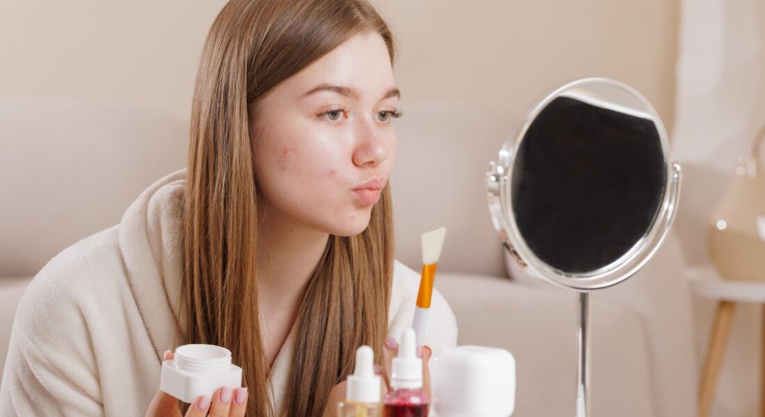 devojka se fleda u ogledalo i drži kremu za lice u ruci