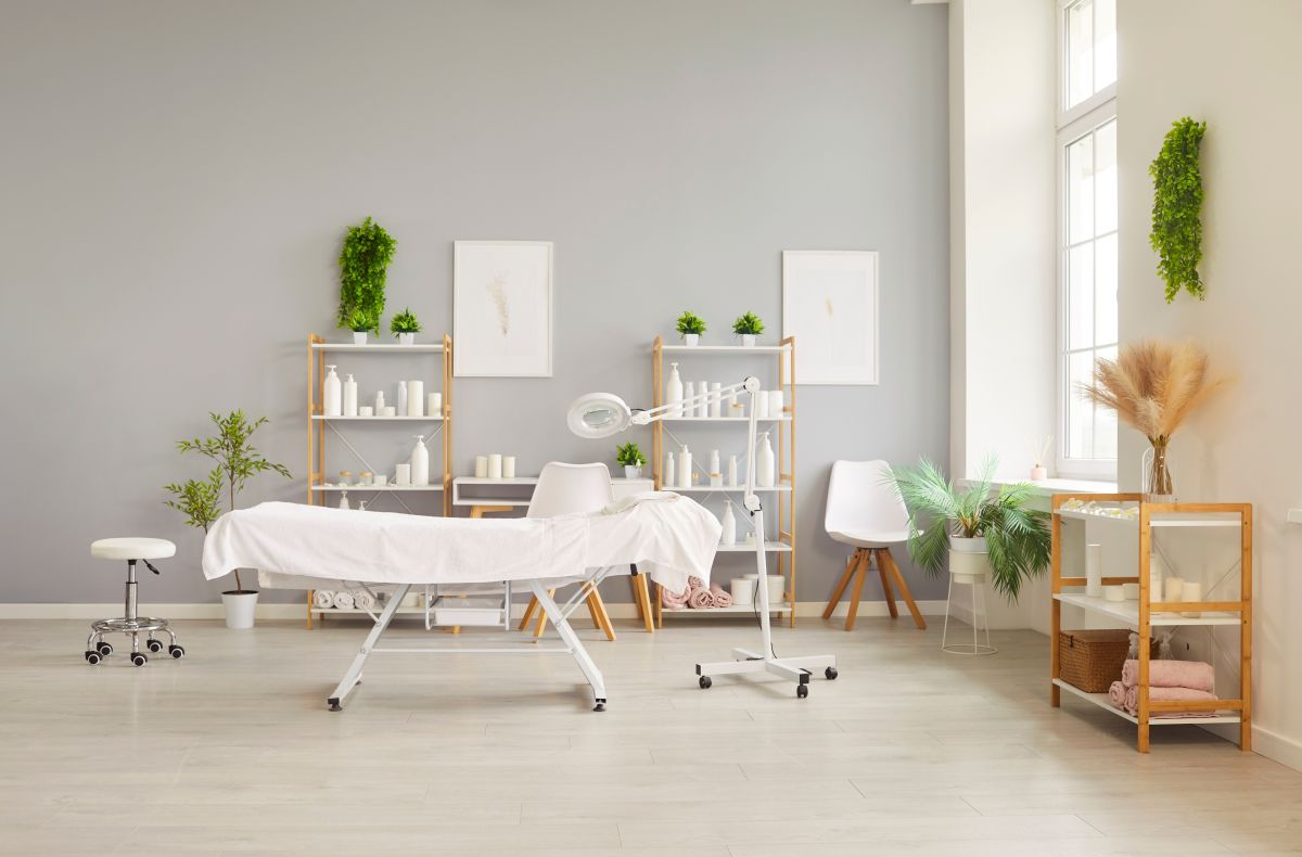Svetao kozmetički salon sa stolom za tretmane, lampom, policama sa preparatima, peškirima i dekorativnim biljkama.